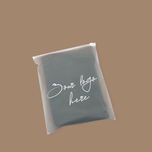Op de afbeelding: Een rechthoekige, halfdoorzichtige opbergzak van mat plastic met een witte ritssluiting aan de bovenkant. De voorzijde van de zak is voorzien van de witte cursieve tekst "Your logo here" in het midden, met een klein rond ventilatiegaatje.