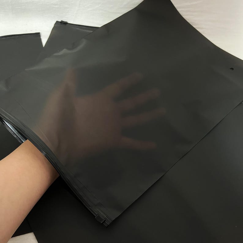 Peut inclure: Plusieurs sacs en plastique noirs, opaques et refermables sont pr&eacute;sent&eacute;s. L'un des sacs est partiellement ouvert, r&eacute;v&eacute;lant une main &agrave; l'int&eacute;rieur. Les sacs ont une fermeture &eacute;clair et semblent &ecirc;tre faits d'un mat&eacute;riau lisse et mat. L'arri&egrave;re-plan est de couleur claire et neutre.