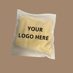 Op de afbeelding: Een doorschijnende, matglazen plastic hersluitbare zak met daarin een opgevouwen geel kledingstuk. De voorzijde van de zak is voorzien van de zwarte tekst "YOUR LOGO HERE" in een vetgedrukt, schreefloos lettertype, gecentreerd op de verpakking.