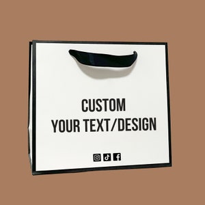 Puede incluir: Bolsa de regalo de papel blanco con ribete negro y asa de cinta negra. La bolsa tiene el texto "CUSTOM YOUR TEXT/DESIGN" impreso en la parte delantera, junto con iconos de redes sociales para Instagram, TikTok y Facebook.