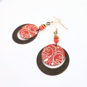 May include: A pair of dangle earrings featuring a circular bronze-toned metal hoop frame. Inside each hoop hangs a smaller, flat, circular disc with a red and white paisley-inspired pattern. A small, round, coral-coloured bead sits above each disc.
