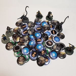 May include: A collection of round, colourful game tokens with various designs, including alien creatures, numbers, and abstract patterns. The tokens are arranged on a white surface.