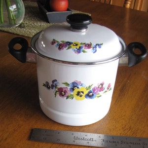 May include: White enamel cooking pot with a lid and black handles. The pot is decorated with a floral pattern of purple, yellow, and blue pansies. A metal ruler is placed next to the pot. The pot measures approximately 7 inches in diameter.