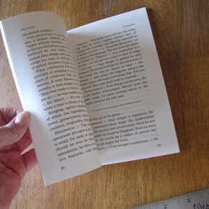 May include: An open book with white pages, held by a hand, revealing text. The book is on a wooden surface with a ruler in the corner. The text is in English, with the word "Stinging" visible.