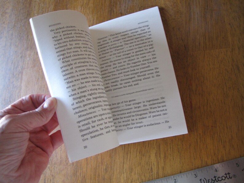 May include: An open book with white pages, held by a hand, revealing text. The book is on a wooden surface with a ruler in the corner. The text is in English, with the word "Stinging" visible.