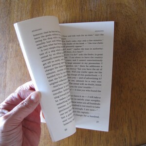 May include: A book with pages being turned, revealing printed text. The pages are off-white, and the text is in black. The book is held by a hand, with the background showing a wooden surface.