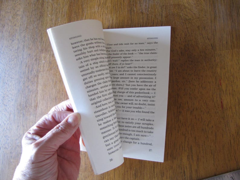 May include: A book with pages being turned, revealing printed text. The pages are off-white, and the text is in black. The book is held by a hand, with the background showing a wooden surface.