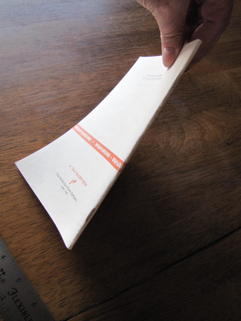 May include: A white paper book with the text "A TALE BLAZER BOOK" and "WRAPAROUND" in orange. The book is held by a person's hand. The book is on a wooden surface.