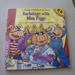 May include: A Muppet Lift-the-Flap Book titled "Backstage with Miss Piggy" featuring Miss Piggy, a pink pig, with blonde hair, wearing a blue robe and purple gloves, getting ready for a performance. Two other pigs, one wearing a red and white striped shirt and the other wearing a blue and white striped shirt, are helping her.