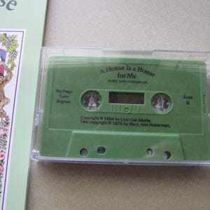 May include: A green cassette tape in a clear plastic case. The tape is labeled "A House is a House for Me" by Mary Ann Hoberman. The cassette is next to a book with colorful illustrations.
