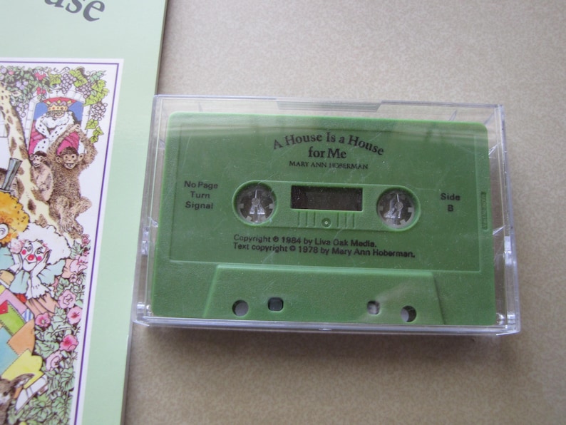 May include: A green cassette tape in a clear plastic case. The tape is labeled "A House is a House for Me" by Mary Ann Hoberman. The cassette is next to a book with colorful illustrations.