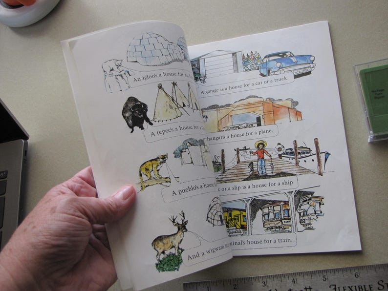 May include: An open book with colorful illustrations and text about different types of houses. The illustrations include an igloo, teepee, garage, hangar, pueblo, slip, and wigwam. The book is being held open by a person.