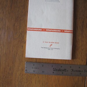 May include: A vintage book titled "A Tale Blazer Book" with a red and white cover, lying on a wooden surface. A flexible ruler is next to the book. The book's cover has the text "WRAPAROUND" and the name of the publisher.