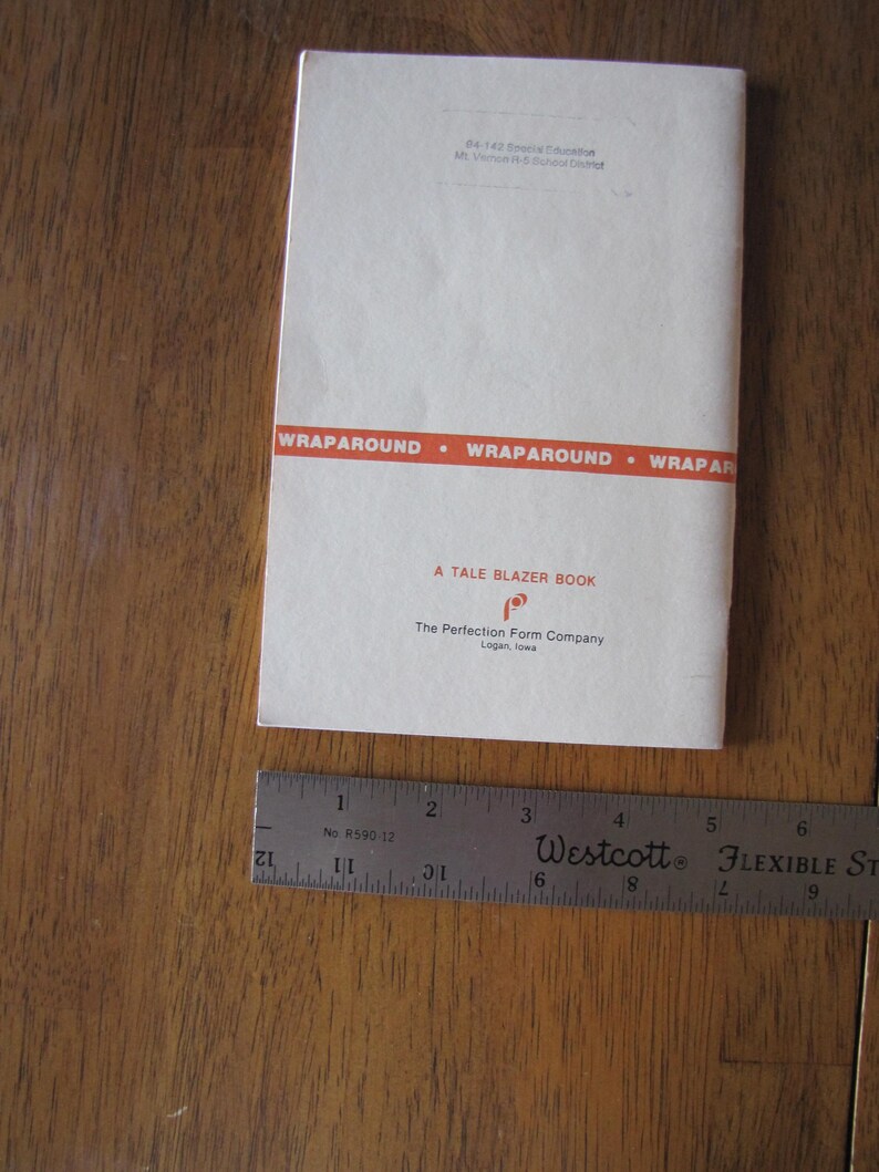 May include: A vintage book titled "A Tale Blazer Book" with a red and white cover, lying on a wooden surface. A flexible ruler is next to the book. The book's cover has the text "WRAPAROUND" and the name of the publisher.