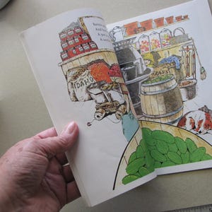 May include: An open book with a colorful illustration of a general store. The illustration features shelves of canned goods, barrels, and a dog sleeping on the floor. A basket of green cucumbers is in the foreground.