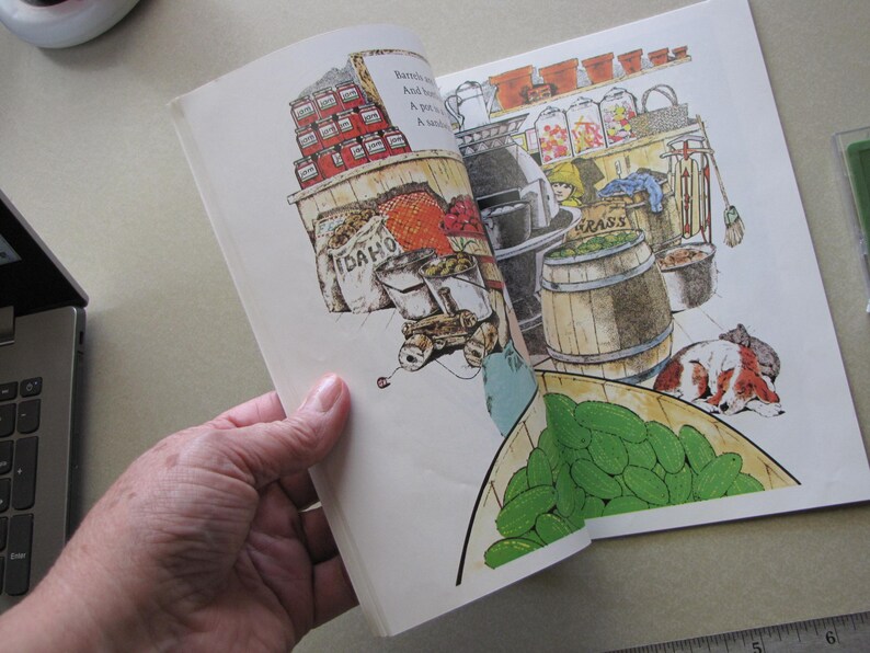 May include: An open book with a colorful illustration of a general store. The illustration features shelves of canned goods, barrels, and a dog sleeping on the floor. A basket of green cucumbers is in the foreground.