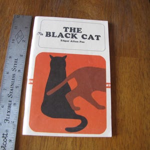 May include: A vintage paperback book titled "The Black Cat" by Edgar Allan Poe. The cover features a black cat silhouette and a brown cat silhouette on an orange background. A metal ruler is next to the book.