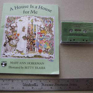 May include: A children's book titled "A House Is a House for Me" with colorful illustrations, next to a green cassette tape in a clear case. A ruler is placed below the book and tape. The book's cover features a detailed drawing of a house and various characters.