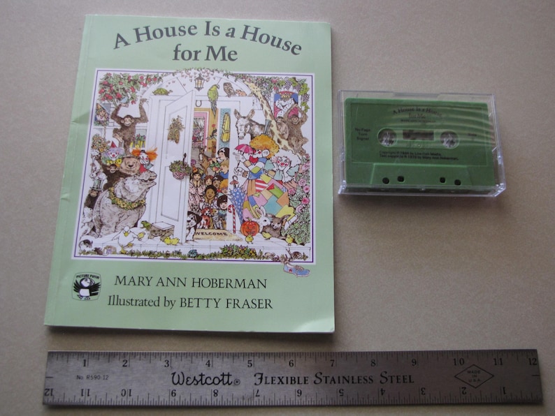 May include: A children's book titled "A House Is a House for Me" with colorful illustrations, next to a green cassette tape in a clear case. A ruler is placed below the book and tape. The book's cover features a detailed drawing of a house and various characters.