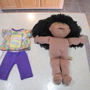 May include: A Cabbage Patch Kid doll with brown skin and black hair wearing a purple outfit. The doll is wearing a yellow and purple floral shirt, purple pants, and a white diaper.