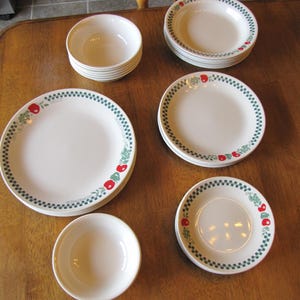May include: A collection of off-white ceramic dinnerware featuring a green checkered border and red apple motifs. The set includes stacks of dinner plates, salad plates, and bowls, all decorated with a consistent pattern of apples and green leaves.