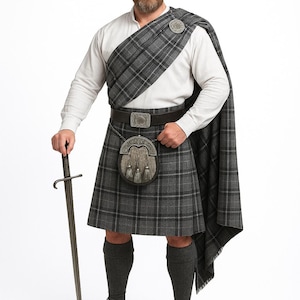 Può includere: Un tradizionale outfit scozzese con kilt nei toni del grigio. L'ensemble comprende un kilt, un plaid abbinato, uno sporran e una lunga spada. L'outfit è completato da una camicia bianca, cintura e calzini grigio scuro. Il kilt presenta un classico motivo tartan.