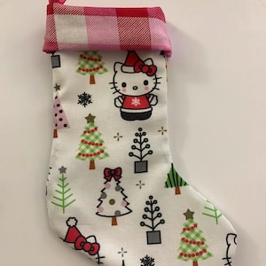 May include: A white Christmas stocking with a pink and red plaid cuff. The stocking features a repeating pattern of green Christmas trees and a Hello Kitty character wearing a Santa hat.