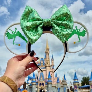 May include: Mouse ear headband featuring a large, green sequined bow. The white ears have a green fairy silhouette. The headband is black and white. A castle and blue sky are in the background.