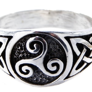 Puede incluir: Un anillo de plata que presenta un medallón circular central con un símbolo de triskel en relieve sobre un fondo oscuro y texturizado. La banda está decorada con patrones de nudos de plata repetitivos a ambos lados del diseño circular.