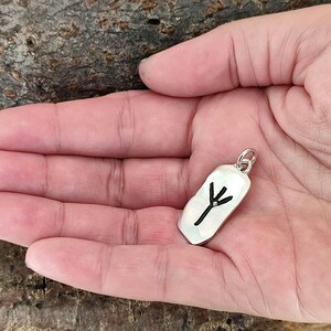 Puede incluir: Un colgante de plata con un s&iacute;mbolo de &aacute;rbol negro grabado en &eacute;l. El colgante est&aacute; en la palma de una mano.