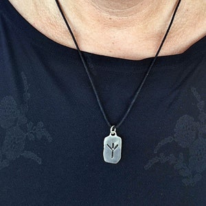 Puede incluir: Un collar con un colgante de plata en un cord&oacute;n negro. El colgante es una etiqueta rectangular con un s&iacute;mbolo de runa grabado.