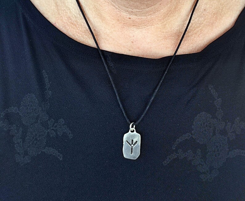 Puede incluir: Un collar con un colgante de plata en un cord&oacute;n negro. El colgante es una etiqueta rectangular con un s&iacute;mbolo de runa grabado.