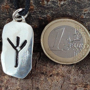 Puede incluir: Colgante de plata con un cord&oacute;n negro. El colgante es un rect&aacute;ngulo de plata con un s&iacute;mbolo de runa negro en su interior. El s&iacute;mbolo de runa es una l&iacute;nea vertical con tres ramas en la parte superior.