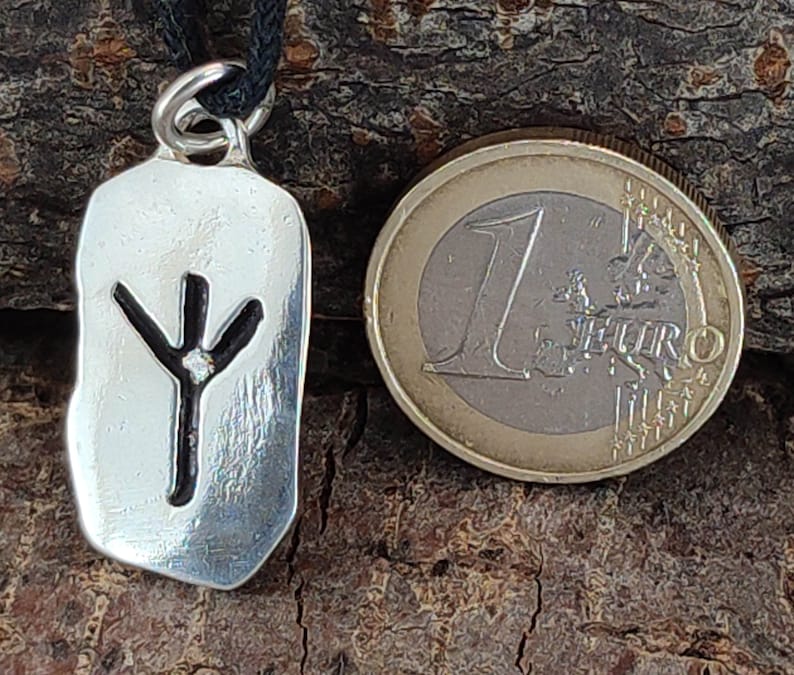 Puede incluir: Colgante de plata con un cord&oacute;n negro. El colgante es un rect&aacute;ngulo de plata con un s&iacute;mbolo de runa negro en su interior. El s&iacute;mbolo de runa es una l&iacute;nea vertical con tres ramas en la parte superior.