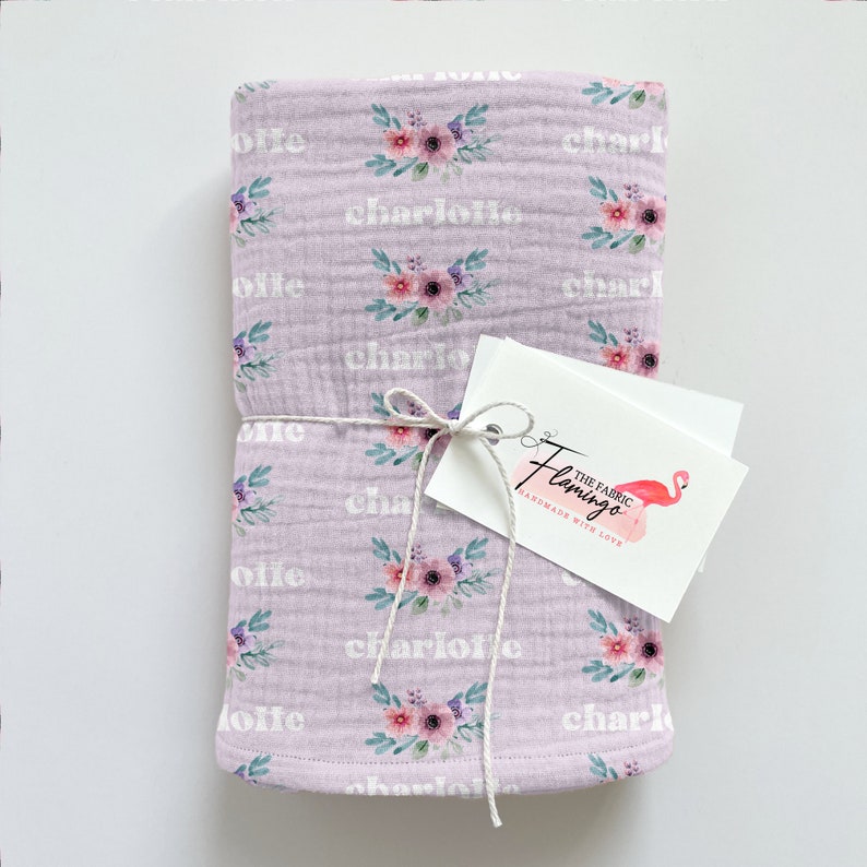 May include: A lavender muslin baby blanket with a floral pattern and the name "Charlotte" repeated throughout. The blanket is tied with twine and a tag that reads "The Fabric Flamingo Handmade with Love".