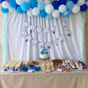 May include: A Frozen-themed birthday party table setting with a blue and white balloon arch, a banner that says "Happy Birthday", and a variety of snacks and desserts. The table is covered in a beige tablecloth.