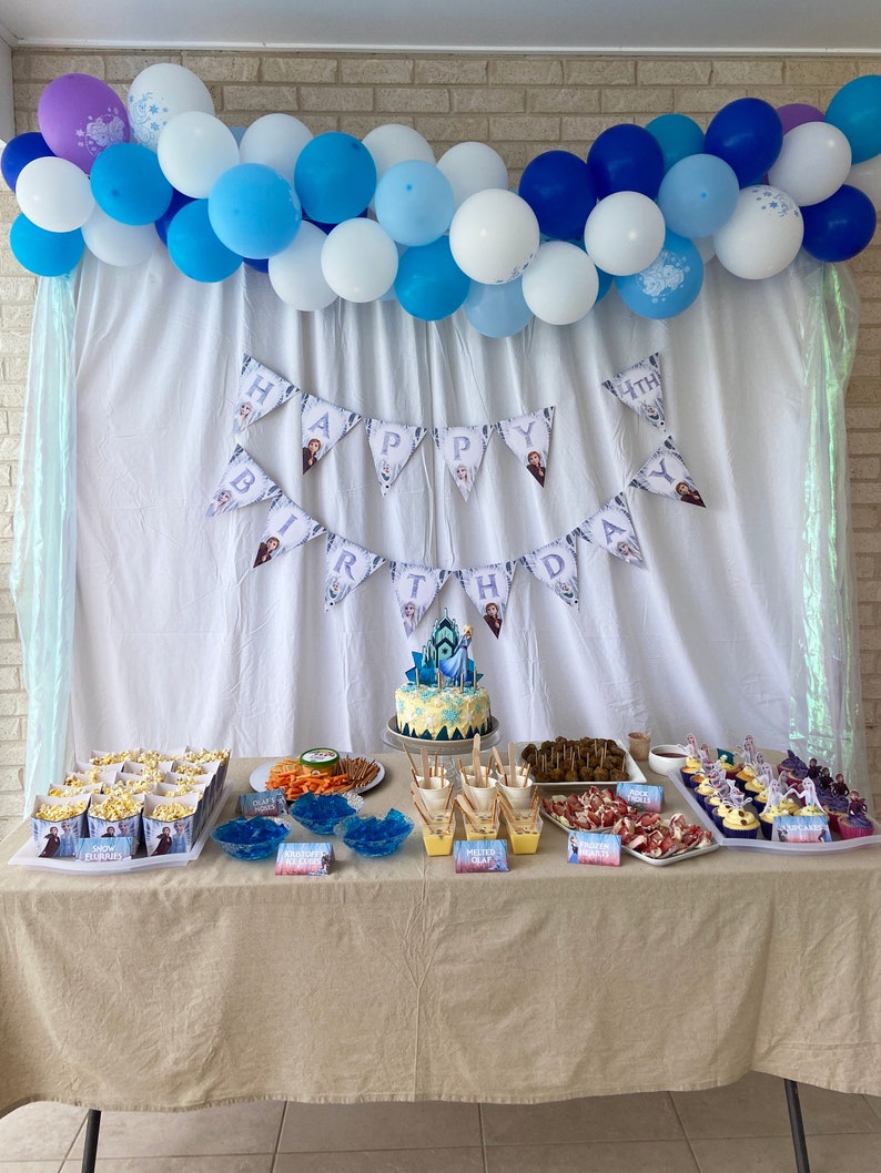 May include: A Frozen-themed birthday party table setting with a blue and white balloon arch, a banner that says "Happy Birthday", and a variety of snacks and desserts. The table is covered in a beige tablecloth.