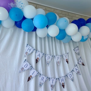 May include: A white backdrop with a blue and white balloon garland. A banner with the words "Happy Birthday" is hanging below the garland.