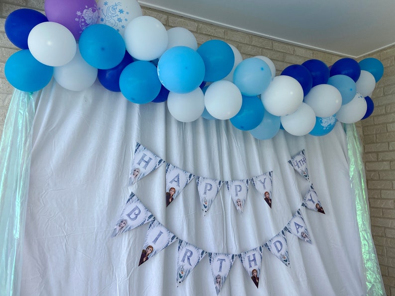 May include: A white backdrop with a blue and white balloon garland. A banner with the words "Happy Birthday" is hanging below the garland.