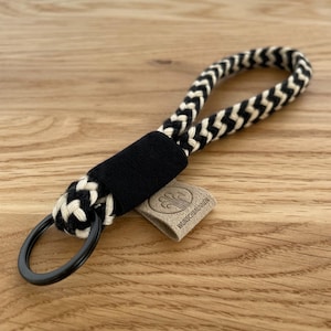 Könnte beinhalten: Schwarz-weiß geflochtener Schlüsselanhänger mit einem schwarzen Metallring und einem braunen Lederanhänger mit dem Text "Wunschbrunnen".
