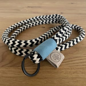 Könnte beinhalten: Schwarzer und weißer Zickzack-Schlüsselband aus Seil mit einem hellblauen Stoffakzent und einem schwarzen Metallring. Der Schlüsselband hat ein braunes Leder-Etikett mit dem Text "The Green Room" in weiß.