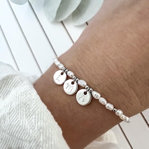 Könnte beinhalten: Ein zartes Perlenarmband mit drei silbernen Scheibenanhängern, die jeweils mit einem Buchstaben graviert sind. Das Armband besteht aus kleinen, unregelmäßigen Perlen, die mit Silberperlen aufgereiht sind. Die Buchstaben auf den Anhängern sind L, M und A.