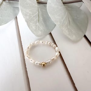 Op de afbeelding: Een delicate ring bestaande uit kleine, gebroken witte parels. Een enkele goudkleurige kraal is gecentreerd op de ring. De ring staat tegen een witte achtergrond, met zachte groene bladeren in het bovenste deel van de afbeelding.