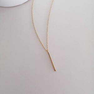 Puede incluir: Un collar de cadena dorada con un colgante de barra vertical delgada.