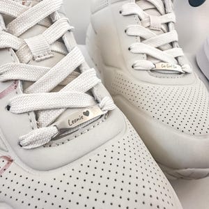 Könnte beinhalten: Nahaufnahme von zwei weißen Turnschuhen mit weißen Schnürsenkeln und perforierten Details. Jeder Schuh hat ein Metallschild mit Text: Einer sagt "Leonie" mit einem Herz, der andere sagt "Go Girl Go".