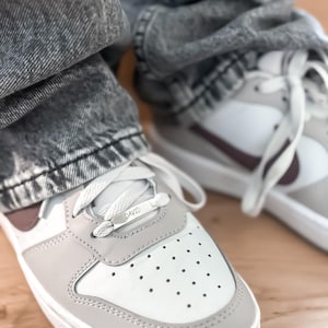 May include: Close-up of a pair of white and gray sneakers with a burgundy swoosh and accents. The sneakers have white laces and the word "DAVID" is on the lace tag. The shoes are paired with gray acid wash jeans.