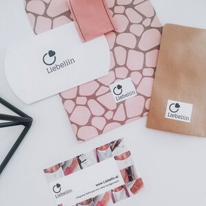 Puede incluir: Un sobre blanco con el logotipo "Liebeliin" y un coraz&oacute;n. El sobre est&aacute; sobre un papel estampado rosa y marr&oacute;n con un cuadrado de tela rosa. Hay un sobre de papel marr&oacute;n con el logotipo "Liebeliin" y un coraz&oacute;n. Una tarjeta de visita con el logotipo "Liebeliin" y un coraz&oacute;n, el sitio web "www.Liebeliin.at" y el texto "Filigraner Schmuck mit Liebe handgemacht" est&aacute; sobre una superficie blanca.