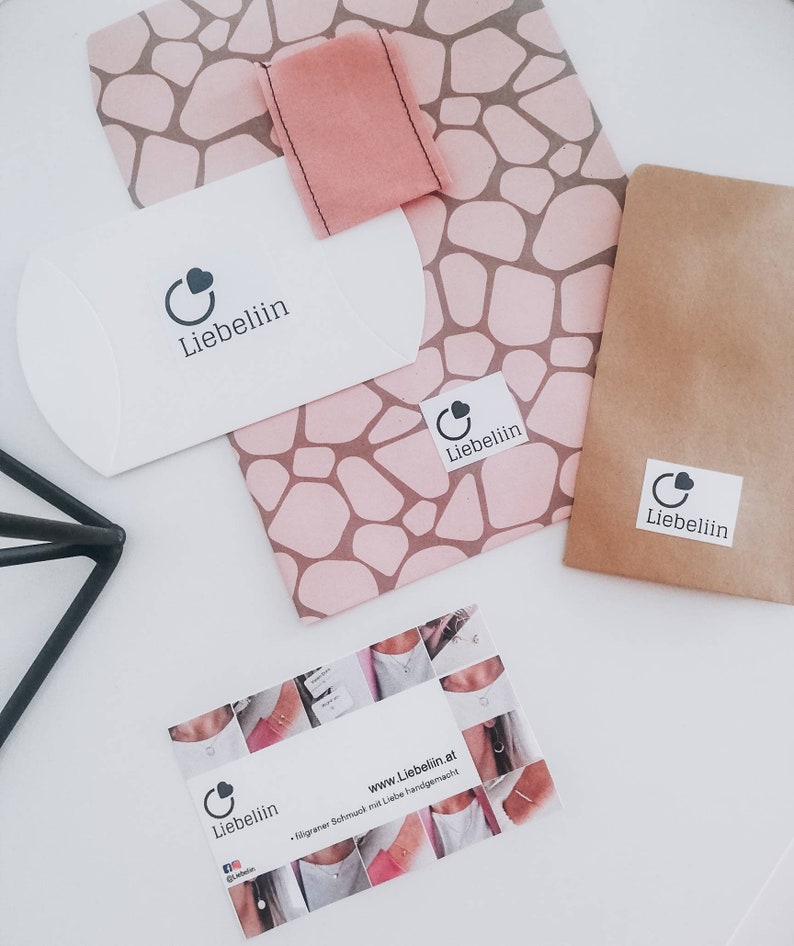 Puede incluir: Un sobre blanco con el logotipo "Liebeliin" y un coraz&oacute;n. El sobre est&aacute; sobre un papel estampado rosa y marr&oacute;n con un cuadrado de tela rosa. Hay un sobre de papel marr&oacute;n con el logotipo "Liebeliin" y un coraz&oacute;n. Una tarjeta de visita con el logotipo "Liebeliin" y un coraz&oacute;n, el sitio web "www.Liebeliin.at" y el texto "Filigraner Schmuck mit Liebe handgemacht" est&aacute; sobre una superficie blanca.