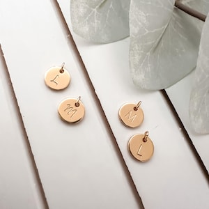 Peut inclure: Quatre petits pendentifs ronds de couleur or avec des lettres gravées : L, M, M et L. Chaque pendentif a une petite boucle en haut pour l'attacher à une chaîne ou un bracelet. Les pendentifs sont disposés sur une surface en bois blanc.