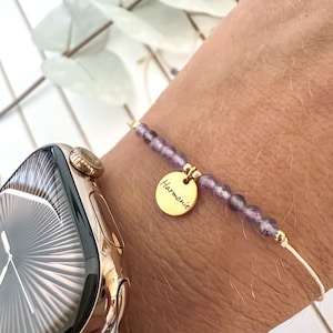 Puede incluir: Una delicada pulsera con pequeñas cuentas redondas moradas y un dije dorado. El dije está inscrito con la palabra "Harmonie". La pulsera está ensartada en un cordón fino y claro y se usa en una muñeca junto a un reloj inteligente dorado.